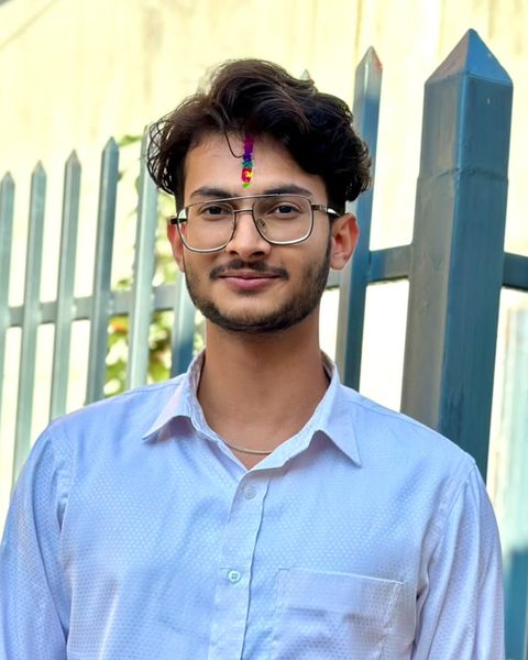 Portrait of Manoj Subedi in a light blue shirt and glasses, standing by a blue iron fence.